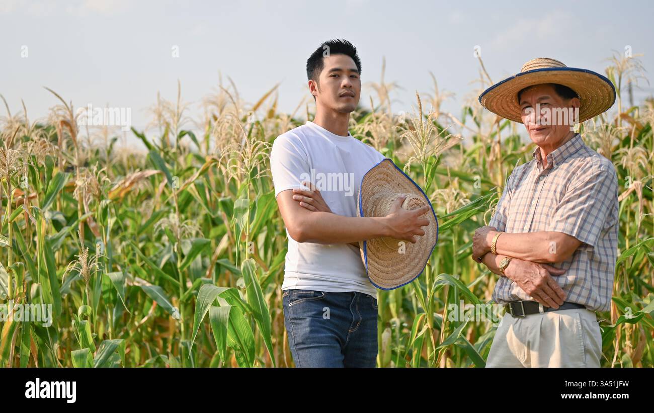 Fiducioso proprietario di una fattoria millenaria asiatica si trova con un vecchio agricoltore in un campo di mais, con le braccia incrociate. L'immagine trasmette la collaborazione nell'agricoltura, nella crescita delle colture e nelle attività rurali. Un'immagine forte per la leadership agricola e le storie di agricoltura a conduzione familiare. Foto Stock