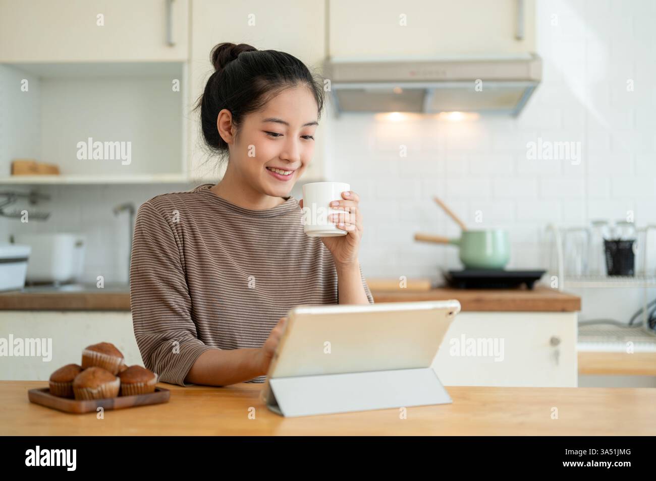 Felice donna asiatica che lavora da casa al tavolo della cucina, sorseggiando un caffè e utilizzando un tablet digitale. Ideale per il lavoro in remoto, l'ufficio domestico e lo stile di vita moderno. Foto Stock