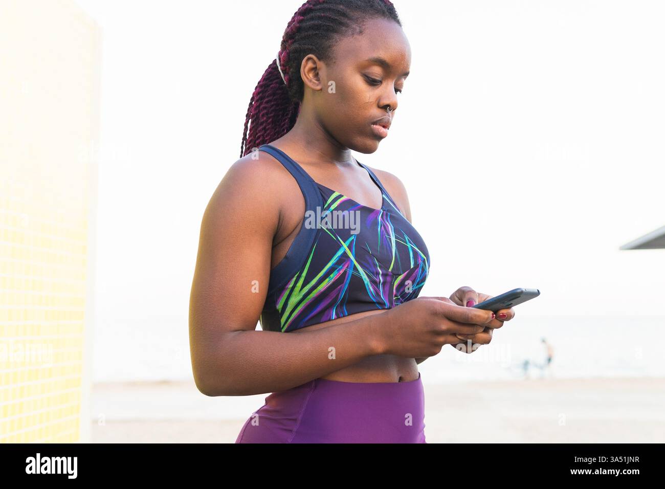 Fit Black atleta femminile in abbigliamento sportivo, si posiziona all'aperto, utilizzando uno smartphone. Sembra concentrata e attiva, perfetta per applicazioni di fitness, campagne di benessere e immagini di tecnologia mobile. Foto Stock