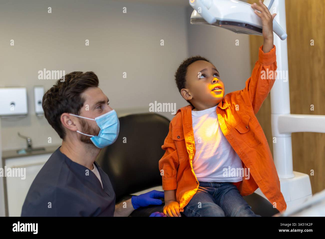 In una clinica odontoiatrica, un ragazzo dalla pelle scura guarda con interesse una lampada dentale. Questo momento sanitario è adatto per odontoiatria pediatrica, scenari medici e pazienti e argomenti di tecnologia dentale. Foto Stock