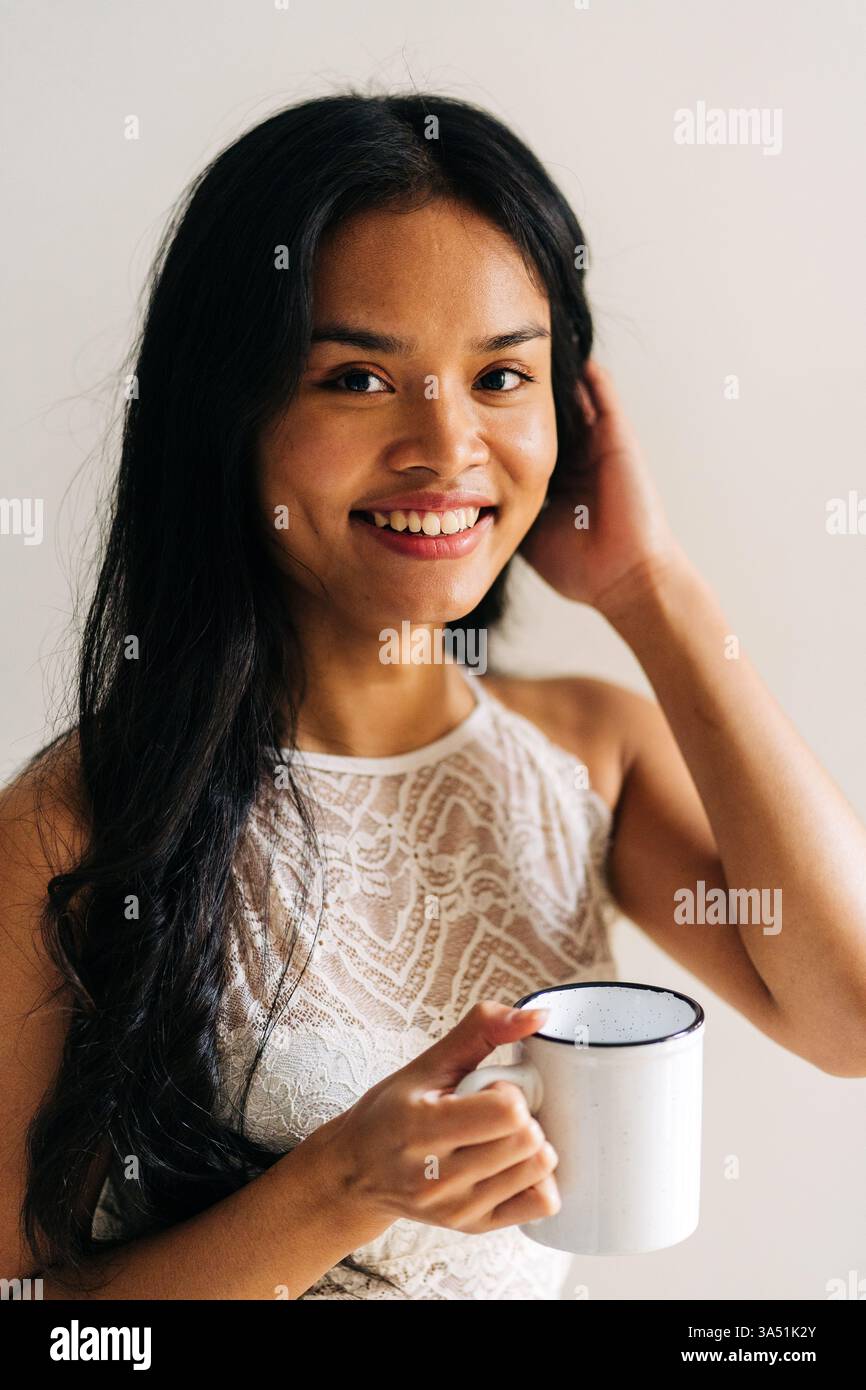 Donna asiatica sorridente che tocca i capelli tenendo una tazza di caffè a casa Foto Stock Donna asiatica sorridente che tocca i capelli tenendo una tazza di caffè a casa Foto Stock