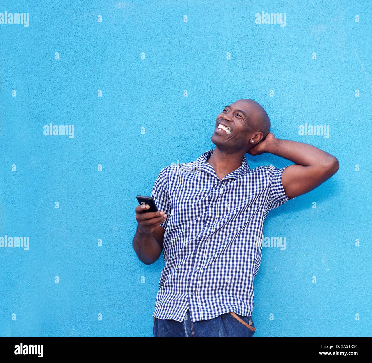 Uomo nero sorridente mano dietro la testa che tiene lo smartphone appoggiato su sfondo blu Foto Stock
