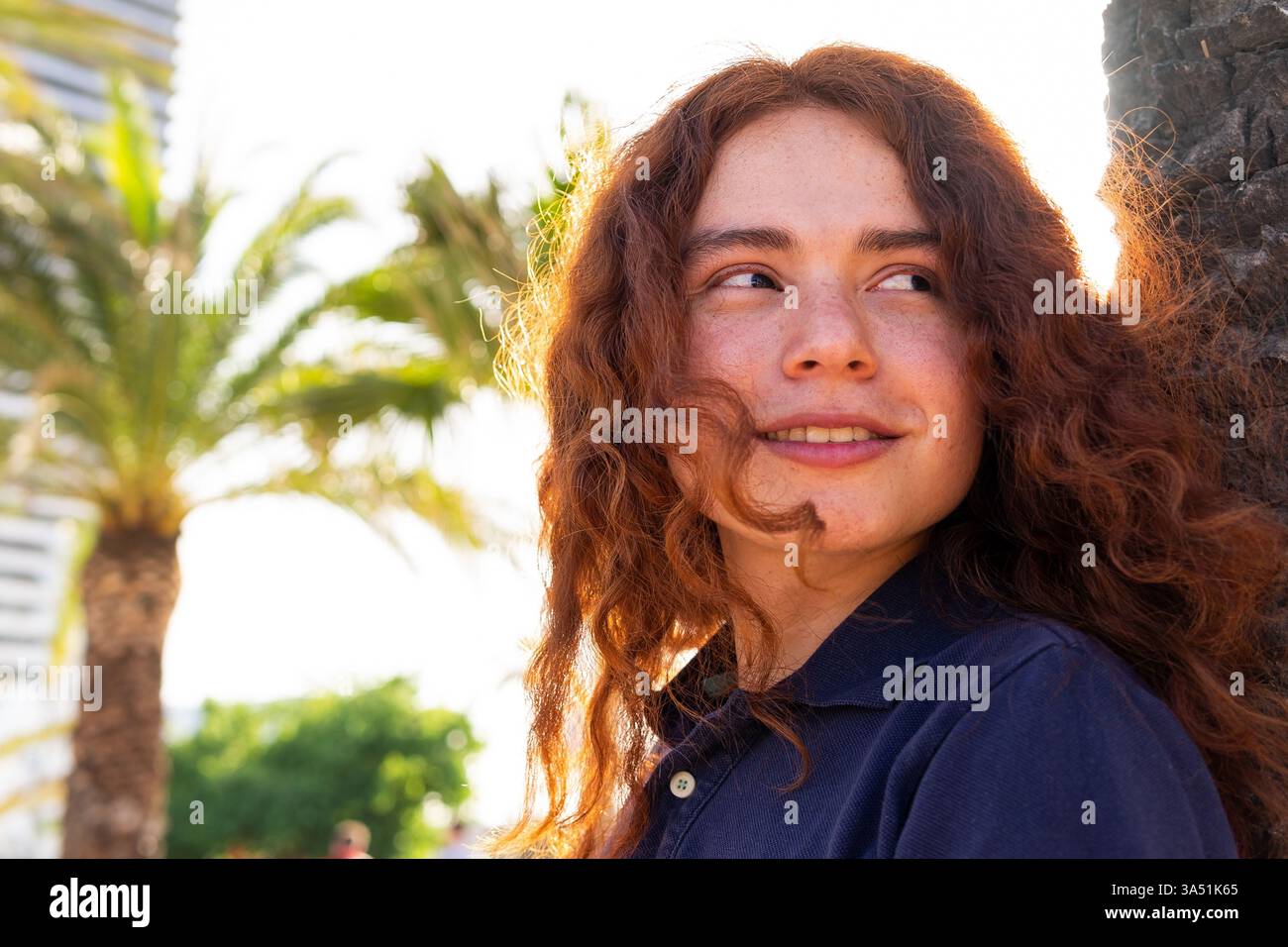 Ritratto in vita di un uomo ispanico sorridente in un parco cittadino fiancheggiato da palme in una giornata di sole. Questa foto lifestyle all'aperto trasmette sicurezza e un'atmosfera urbana rilassata, adatta per la moda o l'uso promozionale. Lo spazio di copia sul lato migliora le opportunità di posizionamento del testo. Foto Stock