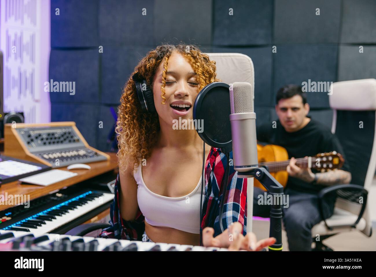 Una donna ispanica canta in un microfono mentre un chitarrista accompagna in uno studio di registrazione. Questa scena da studio musicale è perfetta per registrare, creare talenti e contenuti di intrattenimento. Foto Stock