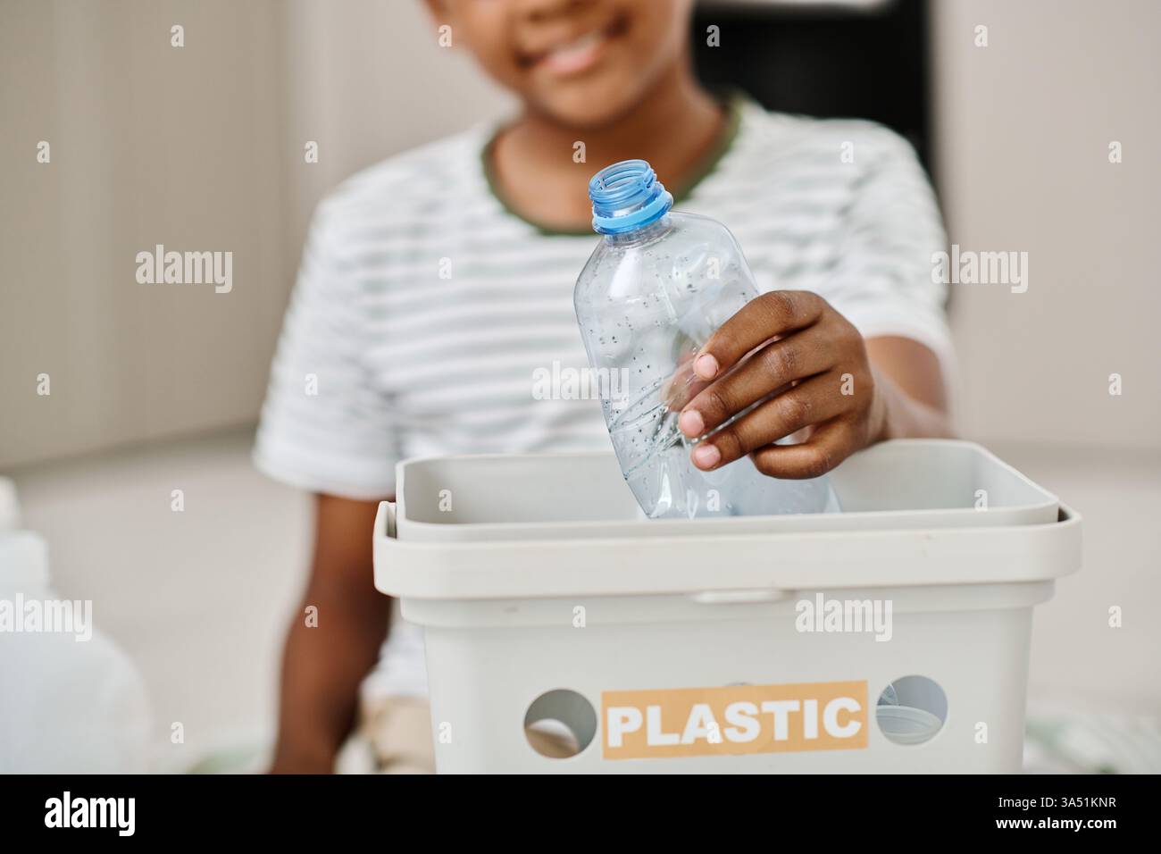 Primo piano di un ragazzo nero che colloca una bottiglia di plastica in un contenitore di riciclaggio. Il momento mette in luce la separazione dei rifiuti e la responsabilità ambientale. Adatto per il riciclaggio, l'ecologia e le campagne scolastiche o familiari. Foto Stock
