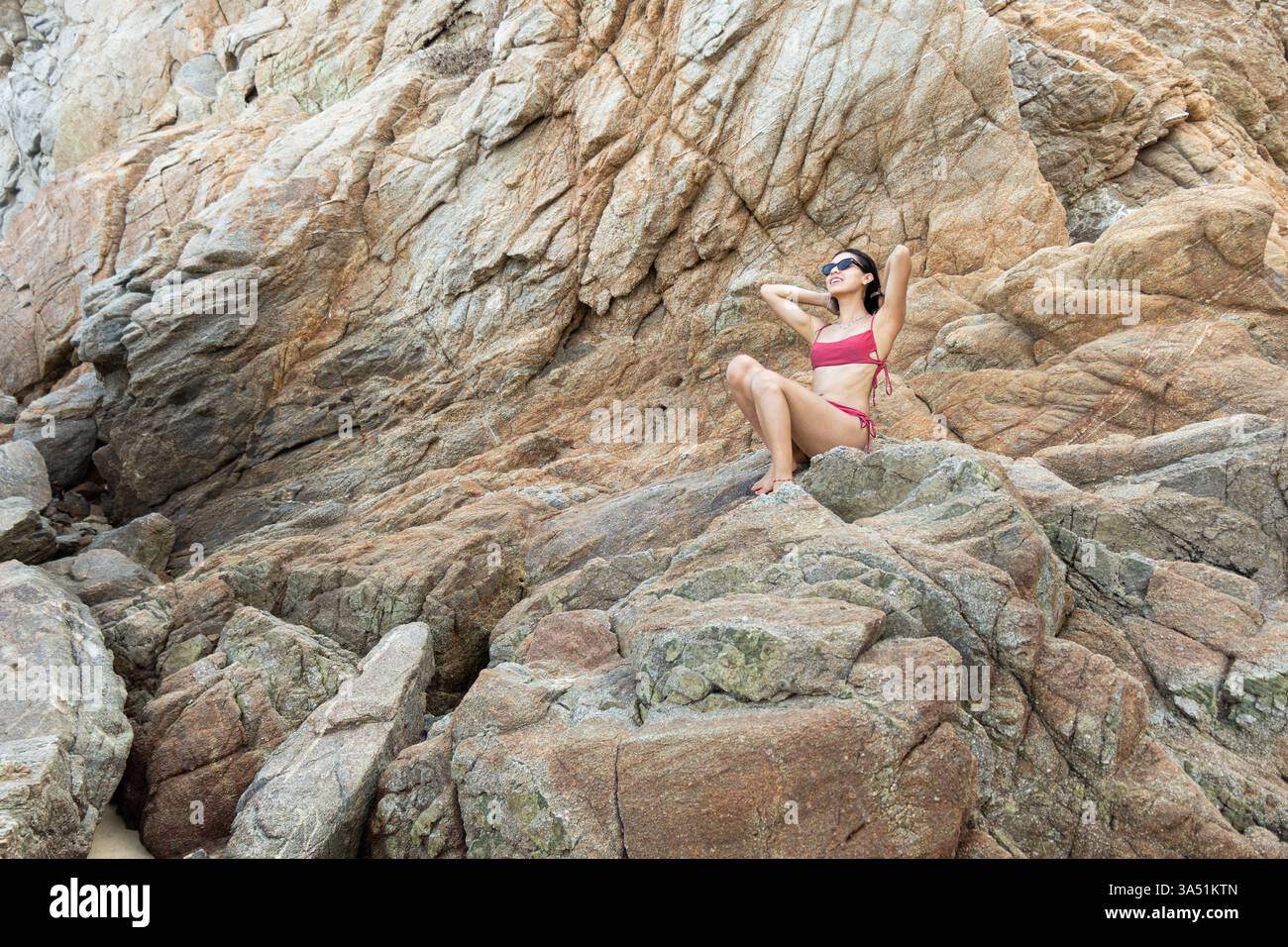 Donna ispanica sorridente in bikini rosa e occhiali da sole fissando i capelli e guardando lontano mentre è seduta su una collina rocciosa durante le vacanze estive Foto Stock