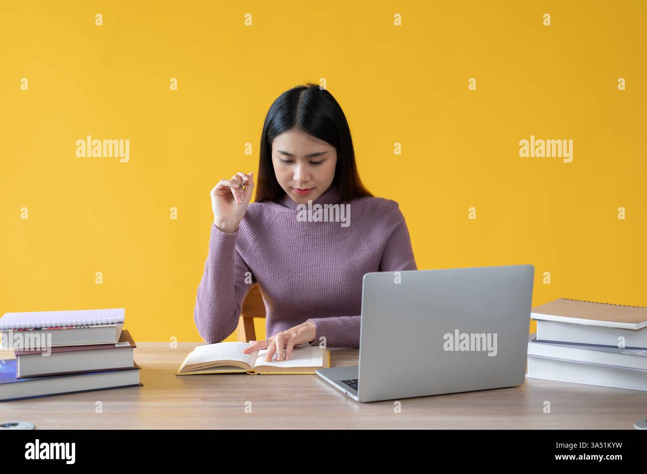 Donna asiatica concentrata che legge un libro alla sua scrivania con uno sfondo giallo brillante. Questa immagine funziona bene per i contenuti educativi, di studio e di sviluppo professionale con studenti o donne d'affari. Ideale per l'ufficio e i temi di apprendimento. Foto Stock