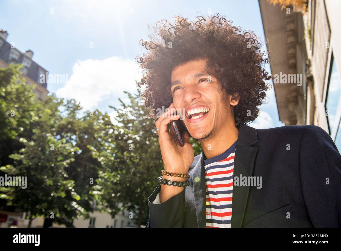 Scena all'aperto con un gioioso afroamericano al telefono. Guarda lontano mentre ride, trasmettendo cordialità e connessione in un ambiente naturale. Un momento di vita sincero adatto a comunicazioni, tecnologia mobile e temi di umore positivo. Foto Stock