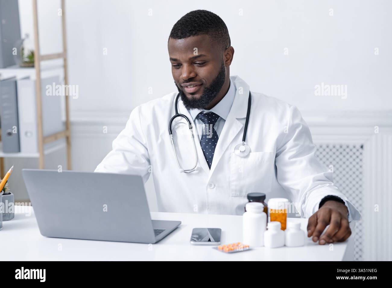 Un medico africano concentrato controlla nuove pillole online, usando un portatile in clinica. Una moderna immagine sanitaria e farmaceutica adatta per la sanità digitale, la telemedicina e le campagne farmacologiche. Perfetto per comunicazioni mediche, marketing in farmacia e ambienti clinici. Foto Stock