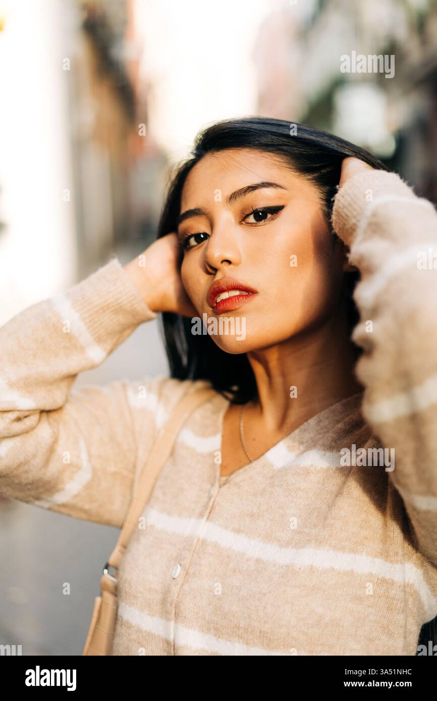 Una giovane donna che indossa un abito morbido con un ornamento a righe guarda la fotocamera alla luce del sole della città. Il ritratto urbano combina stile contemporaneo con luce naturale. Ideale per la moda, lo stile di vita e la fotografia in città. Foto Stock