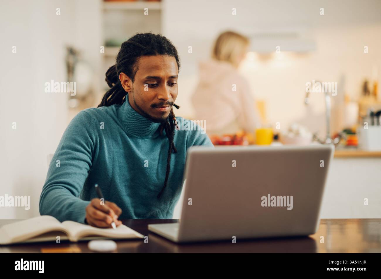 Immagine dello stile di vita di una scena domestica multiculturale. Un assistente online arabo si siede al tavolo da pranzo, scrivendo in un programma mentre sua moglie cucina in cucina. Ideale per contenuti sul lavoro in remoto, il freelance e la gestione della vita lavorativa da casa. Foto Stock