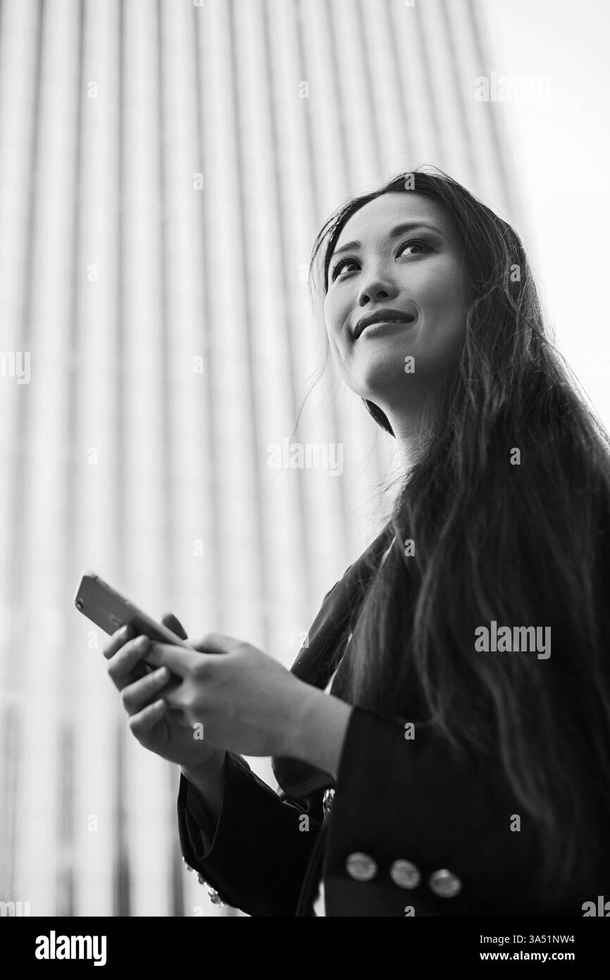 Felice donna asiatica che naviga su un telefono cellulare in una città trafficata. Un'immagine di stile di vita urbano moderno che cattura la vita connessa. Foto Stock