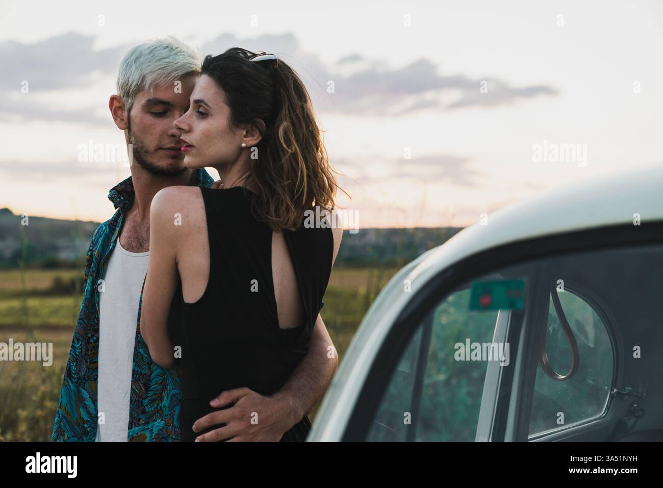 Una coppia in abito elegante abbraccia vicino a un'auto con un paesaggio panoramico sullo sfondo. Ideale per viaggi romantici, stili di vita e immagini di vacanza in campagna o in campagna. Foto Stock