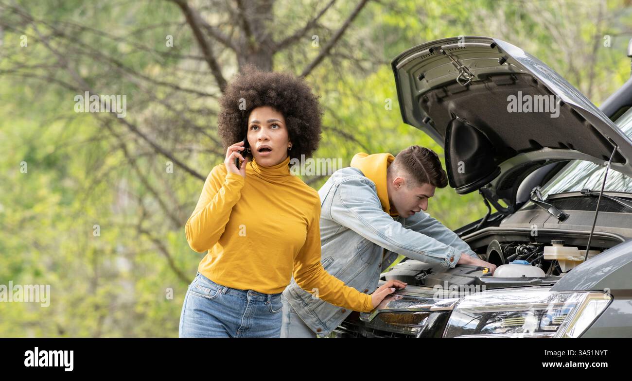 Scena naturale sul ciglio della strada che mostra una donna nera al telefono mentre un uomo bianco ripara un'auto. Questo momento diversificato si adatta all'assistenza automobilistica, all'assistenza stradale e alle campagne di viaggio. Un'immagine diurna contemporanea che mette in risalto la comunicazione e il supporto. Foto Stock