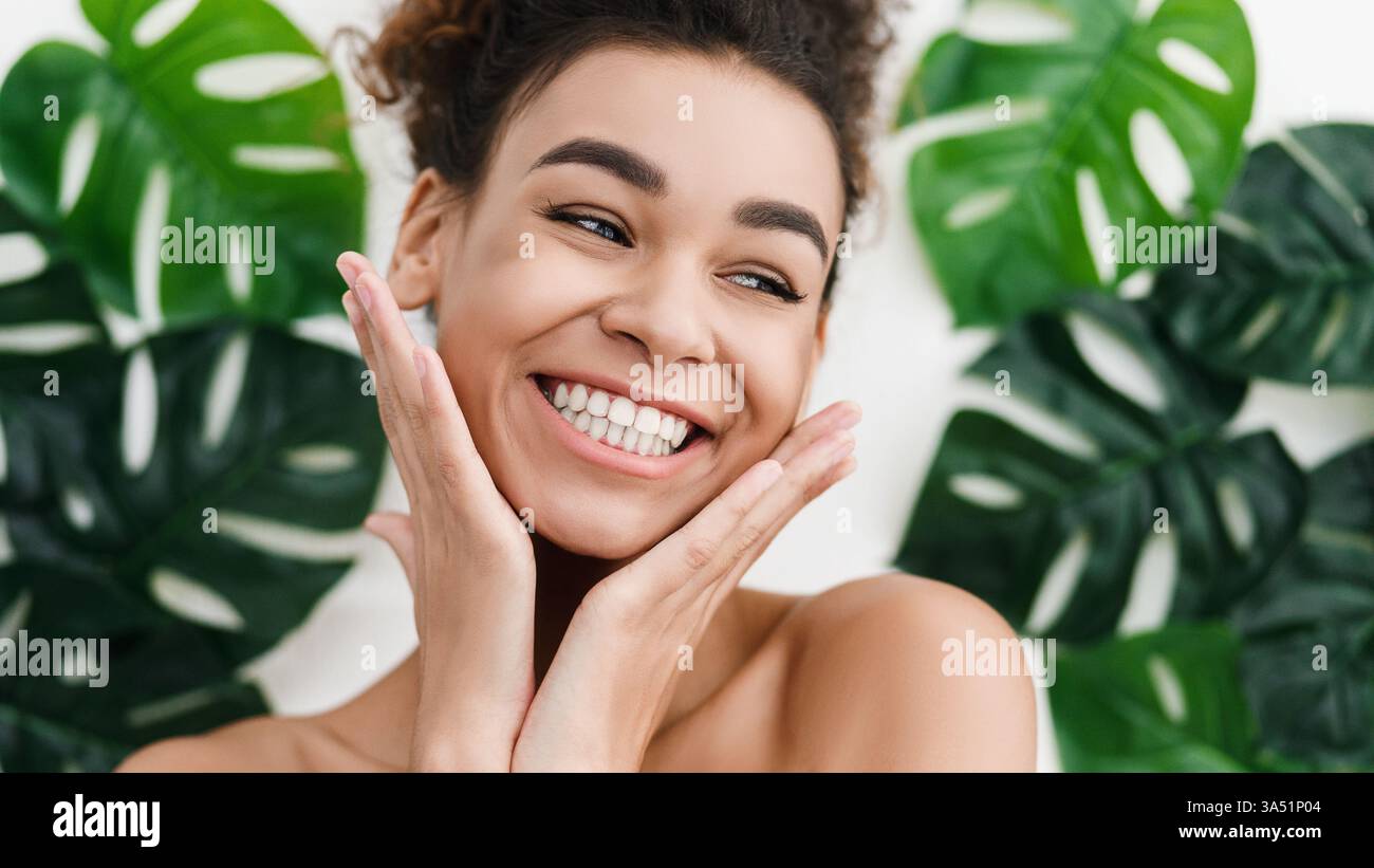Donna nera sorridente che tocca il viso guardando lontano mentre si sta in piedi contro il muro con grandi foglie in studio Foto Stock Donna nera sorridente che tocca il viso guardando lontano mentre si sta in piedi contro il muro con grandi foglie in studio Foto Stock