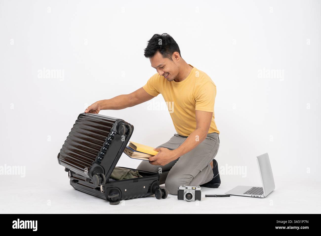 Un uomo asiatico sorridente apre i suoi bagagli e si siede sul pavimento sullo sfondo di uno studio bianco. Questa immagine per la preparazione del viaggio è ideale per lo stile di vita e i temi del confezionamento. Foto Stock