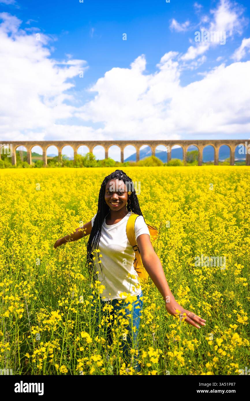 Sorridendo nella natura, una ragazza di etnia nera con trecce, un viaggiatore, in un campo di fiori gialli Foto Stock