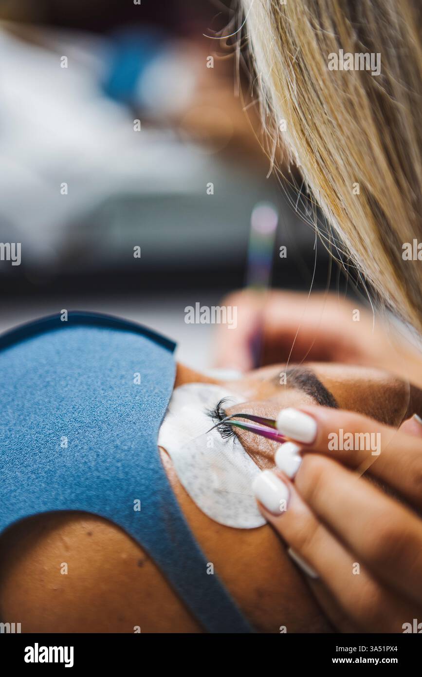 Una donna ispanica che indossa una maschera riceve una procedura di estensione delle ciglia in un salone di bellezza. La scena mette in evidenza un trattamento cosmetico professionale in un ambiente interno pulito. Foto Stock