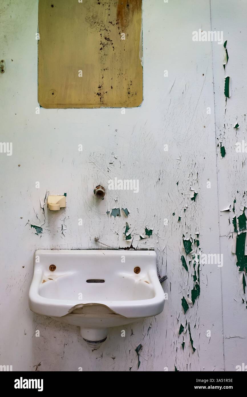 Un lavandino usurato montato su una parete con vernice staccabile e superfici incrinate, all'interno di un bagno abbandonato Foto Stock