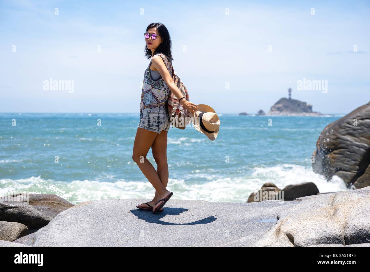 Donna asiatica in piedi su rocce in una giornata di sole, che guarda le spalle. Un'immagine vivace di viaggio e stile di vita adatta alle avventure all'aria aperta, al paesaggio costiero e ai momenti estivi. Ideale per viaggi, esplorazioni e paesaggi panoramici. Foto Stock