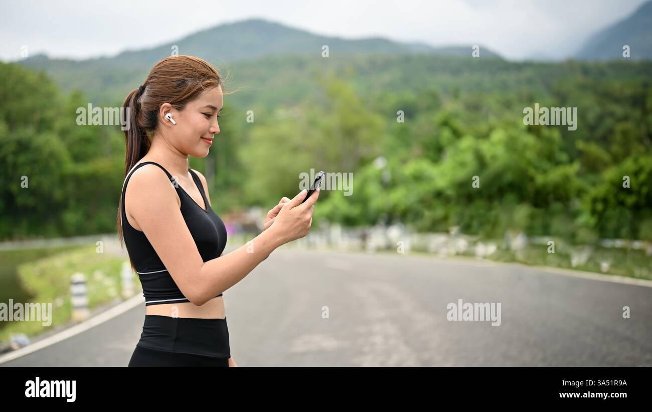 Donna asiatica sorridente con auricolari che utilizzano lo smartphone in piedi per strada durante il giorno Foto Stock