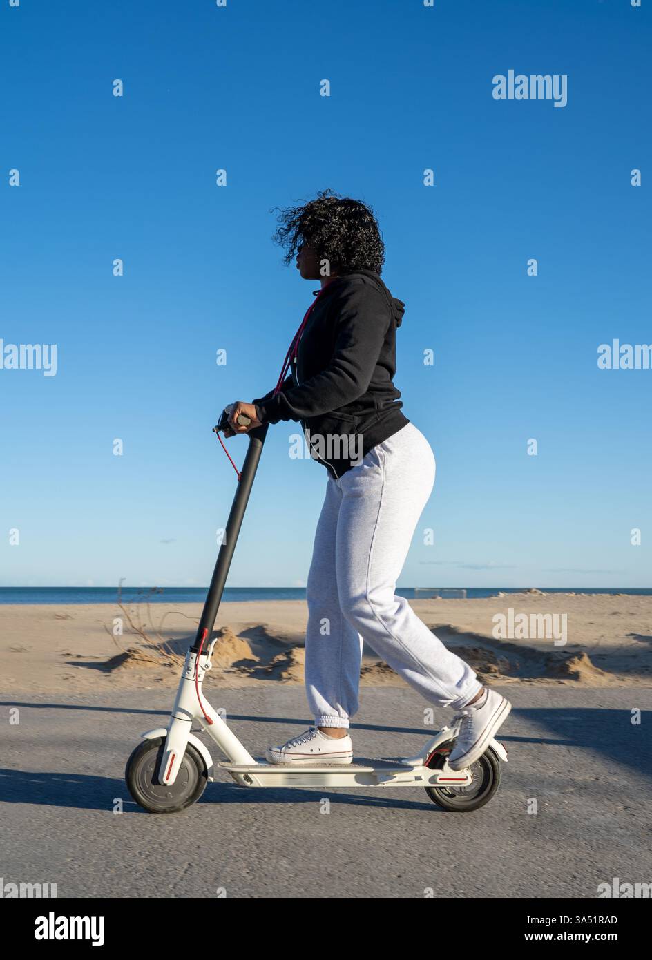 Donna nera che guida uno scooter elettrico lungo una strada asfaltata in riva al mare in una giornata di sole. La scena trasmette mobilità urbana e trasporti ecologici accanto alla spiaggia. Ideale per collezioni di lifestyle, viaggi e trasporti sostenibili. Foto Stock