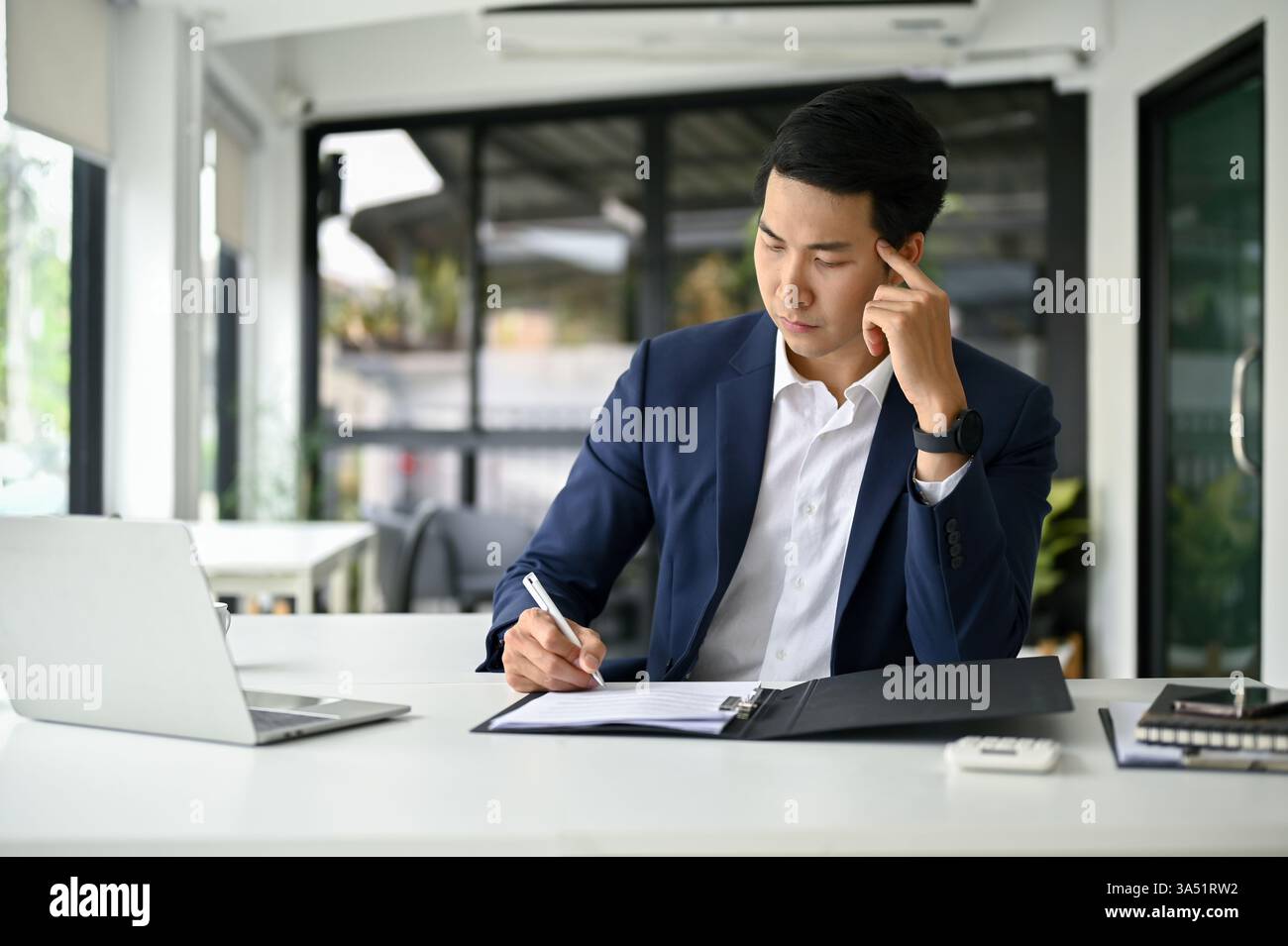 L'uomo d'affari asiatico millenario in una tuta professionale esamina documenti finanziari a una scrivania. La scena trasmette leadership, pianificazione e lavoro aziendale in Thailandia. Ideale per il settore finanziario, aziendale o del branding esecutivo. Foto Stock