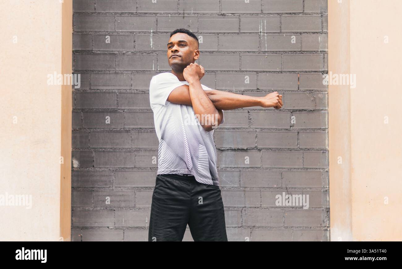 Serio atleta afroamericano maschile in abbigliamento sportivo che si scalda contro un muro di mattoni. L'ambientazione urbana e la concentrazione rendono questa immagine fitness adatta per l'allenamento, la motivazione e il branding sportivo. Foto Stock