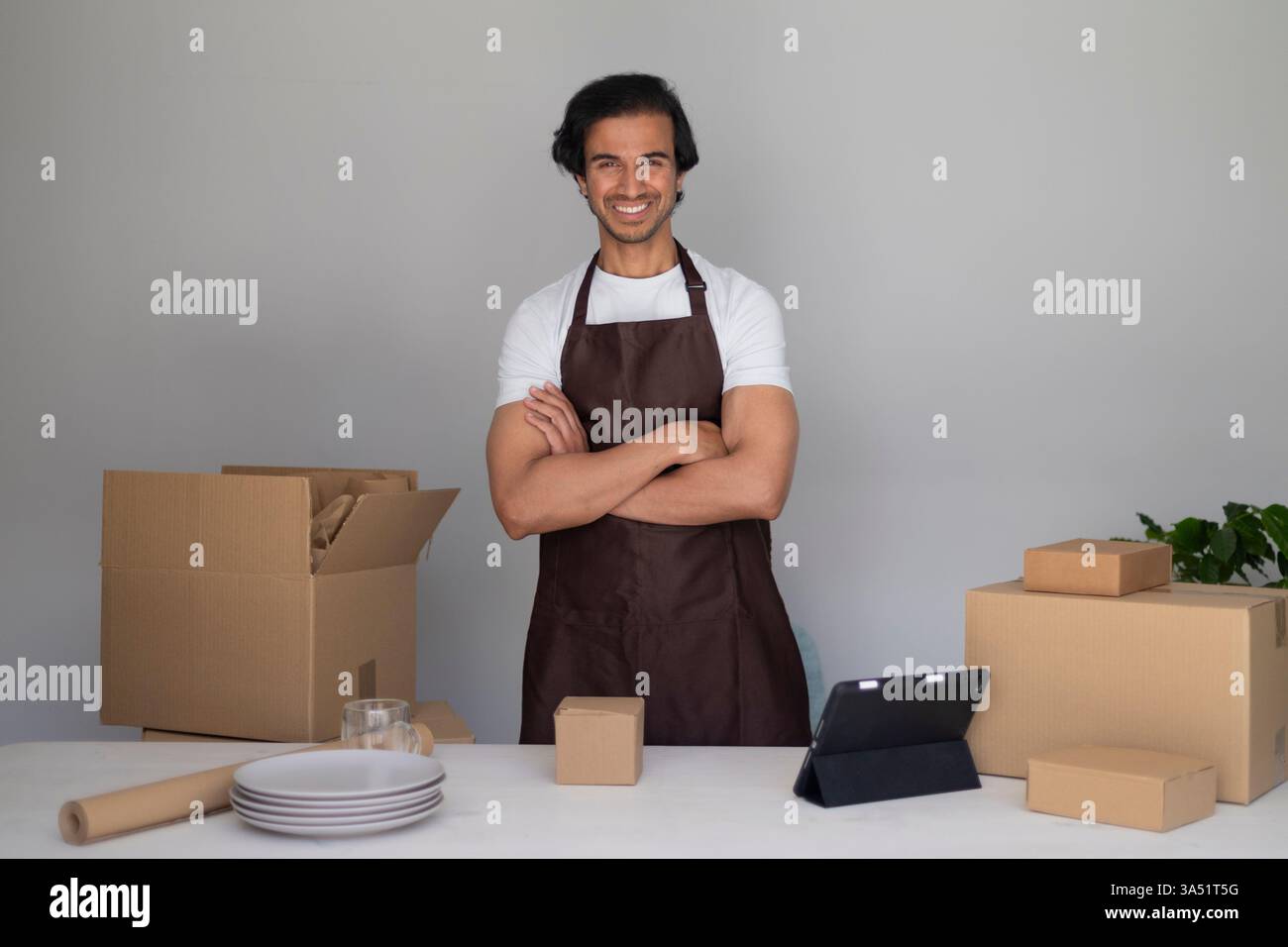 Un uomo sorridente in un grembiule marrone sta con le braccia incrociate in una cucina di casa, circondato da scatole. Questa scena in movimento e disimballaggio trasporta preparazione e stile di vita domestico. Adatto per lo stoccaggio domestico, il trasferimento e la vita familiare. Foto Stock