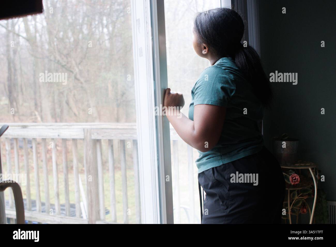 Adolescente nera che guarda fuori dalla finestra del portico posteriore mentre la mano è su vetro Foto Stock