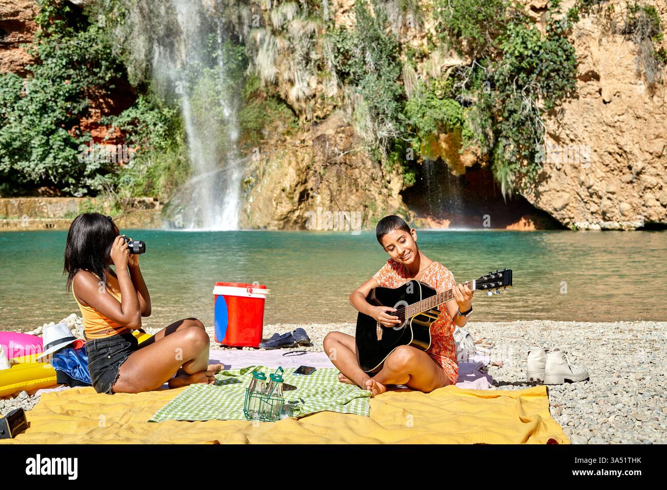Una donna nera fotografa un'amica ispanica che suona la chitarra in una foresta vicino a una cascata in un giorno d'estate. La scena all'aperto enfatizza l'amicizia, la musica e i momenti di viaggio. Ottimo per temi di stile di vita, natura e diversità culturale. Foto Stock