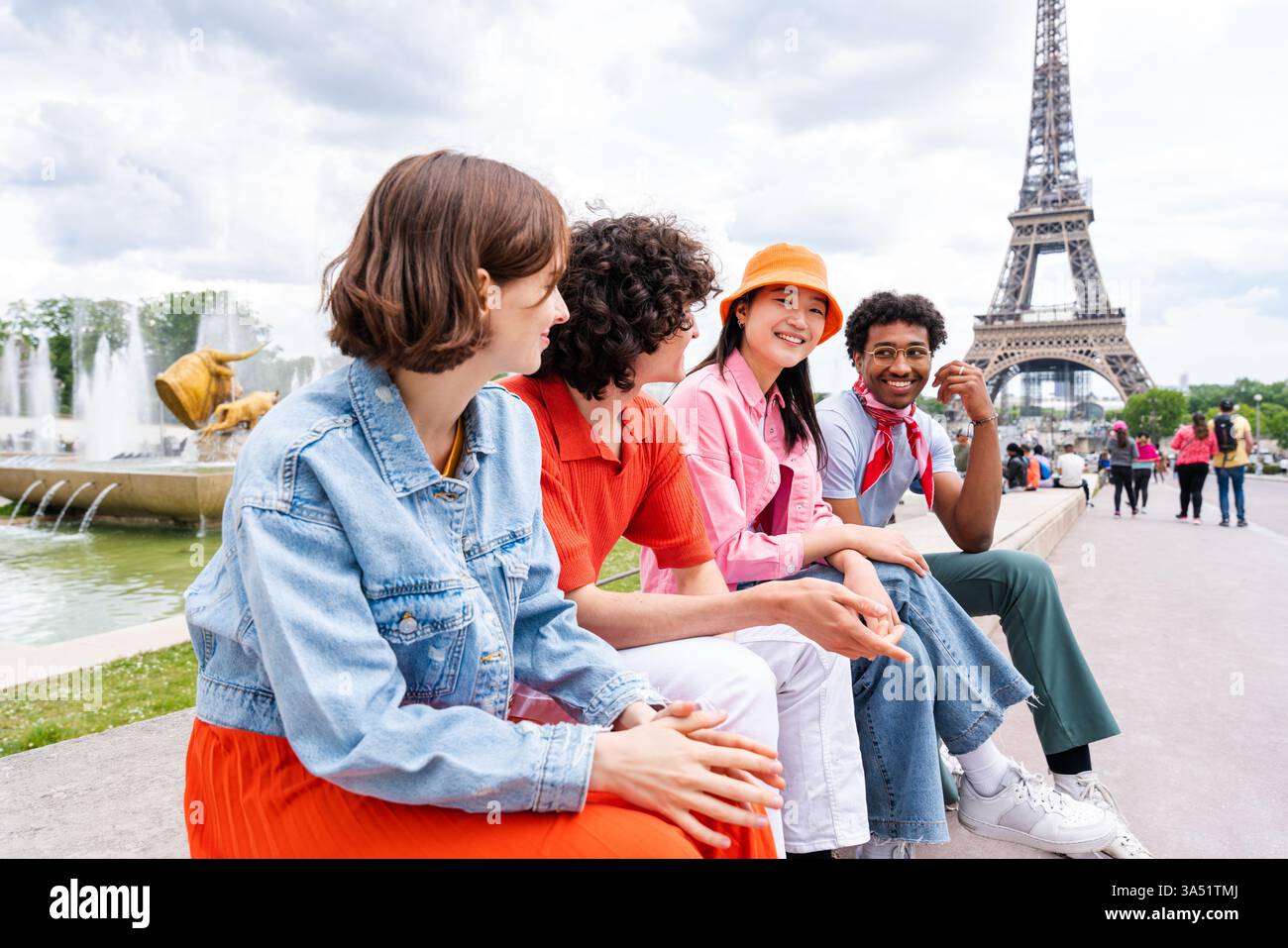 Una donna asiatica sorridente con gli amici siede su una panchina vicino a un punto di riferimento a Parigi, con un cielo nuvoloso sopra la testa. Il momento del viaggio cattura l'amicizia, le visite turistiche e l'esplorazione urbana nella città della luce. Ideale per fare scorta di viaggi, turismo e esperienze di gruppo. Foto Stock