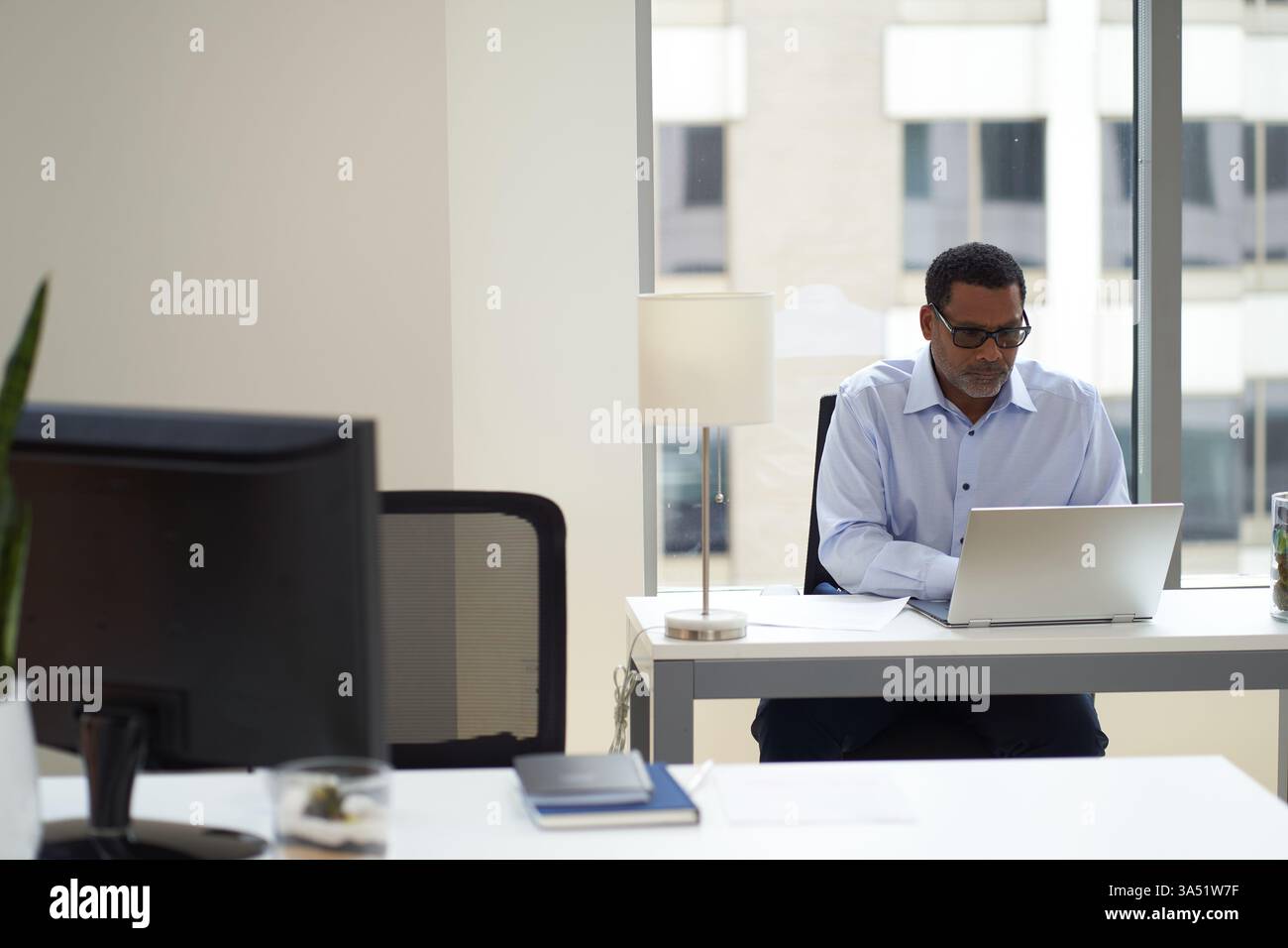 Uomo d'affari afroamericano concentrato in un ufficio intelligente, con occhiali e un laptop. La scena cattura l'attenzione professionale e il lavoro aziendale, adatto a temi aziendali e professionali. Foto Stock