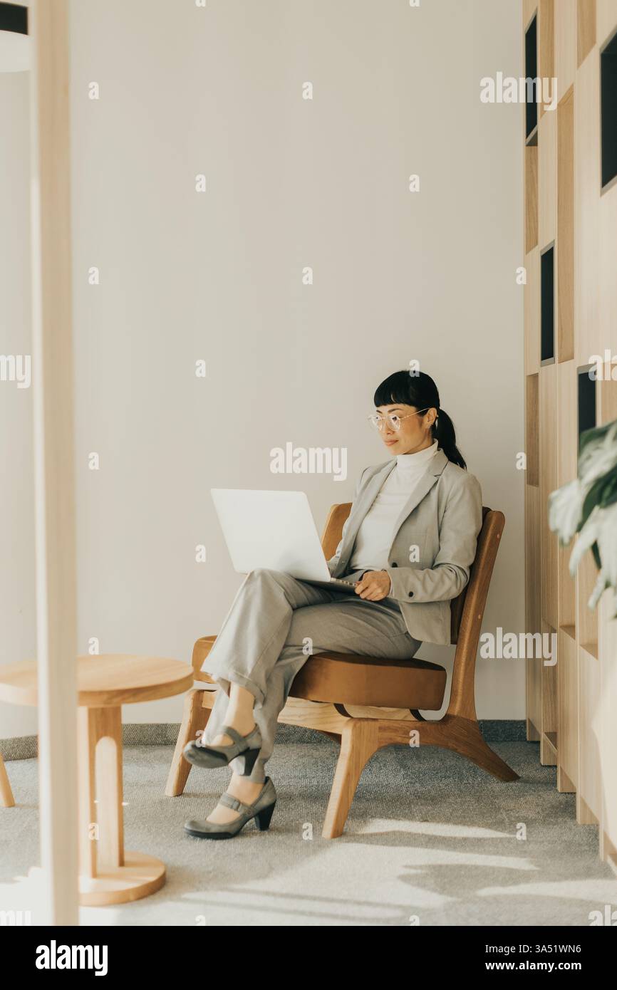 Una donna giapponese professionale e concentrata, vestita con un abito grigio chiaro, si aggancia al proprio computer portatile mentre è seduta comodamente. L'illuminazione soffusa crea un seren Foto Stock