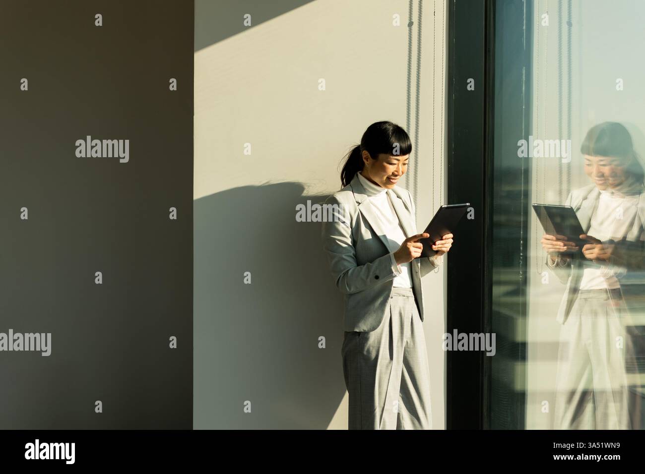 Accanto a una grande finestra in un ufficio moderno, una donna d'affari giapponese usa il suo tablet, assorbito nel suo lavoro. La luce del sole illumina la gioiosa A. Foto Stock