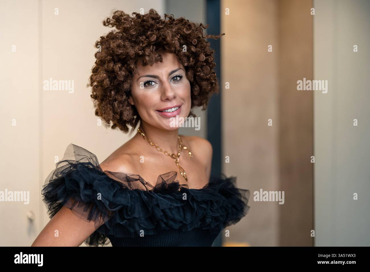 Ritratto di una donna mediorientale sorridente con un taglio di capelli afro che indossa una parte superiore con volant, seduta in casa. L'immagine trasmette un fascino caldo e avvicinabile e casual chic. Foto Stock