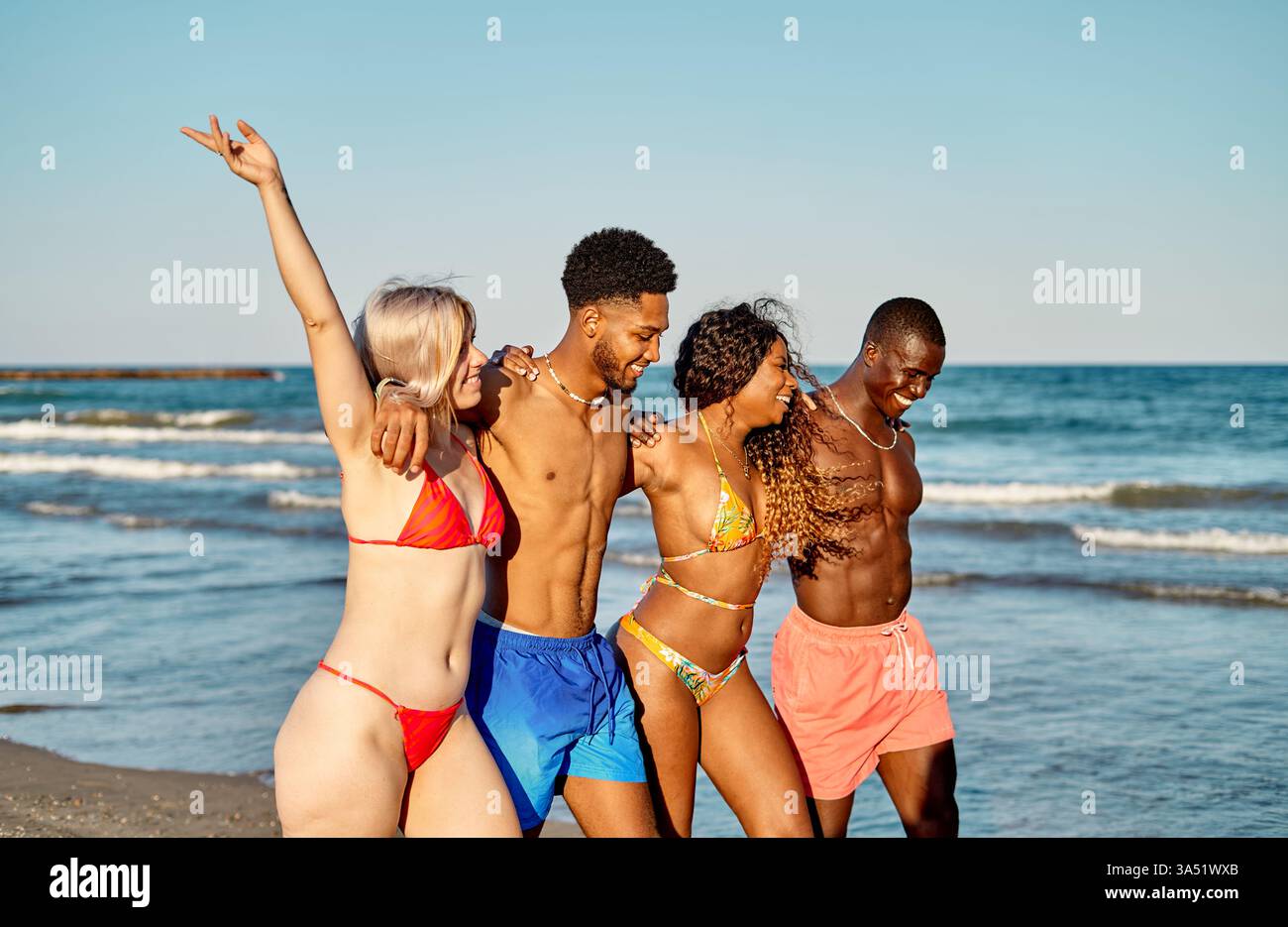 Un gruppo di amici diversi in una passeggiata sulla spiaggia soleggiata, braccia sollevate in un momento celebrativo. Condividono una spensierata giornata estiva in riva al mare, incarnando l'amicizia e le vibrazioni di viaggio. Perfetto per lo stile di vita, i viaggi e il tempo libero all'aperto. Foto Stock