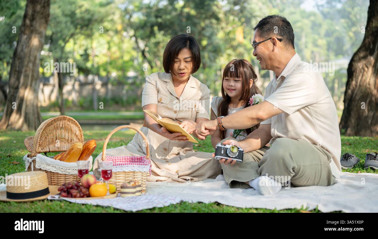 Scena calda di nonni asiatici che leggono una favola alla loro nipote durante un picnic al parco. Condividono un momento di narrazione e di Unione in un ambiente verde all'aperto. Ideale per famiglie, tradizioni e stili di vita all'aperto. Foto Stock