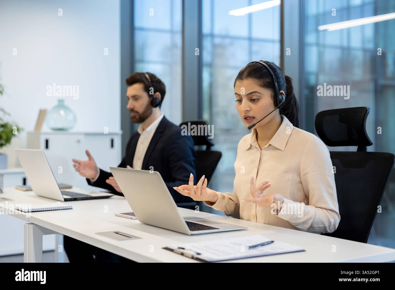 Due impiegati che indossano le cuffie sono impegnati in una videochiamata in un ufficio aperto, dimostrando la comunicazione remota e il servizio clienti. Foto Stock