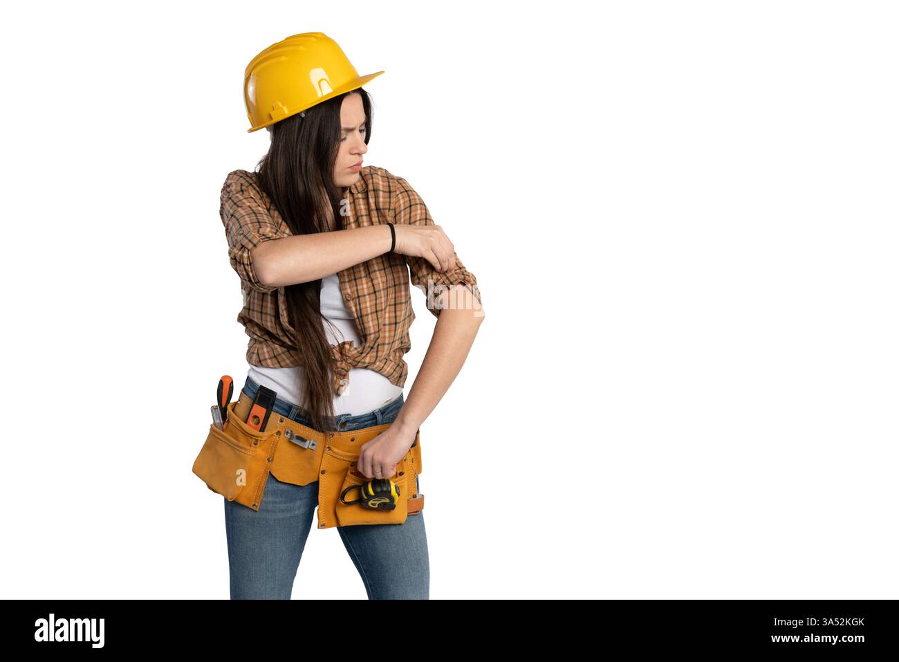 Una lavoratrice edile con un elmetto giallo e una cintura porta attrezzi regola la manica della camicia, preparandosi al lavoro. Ha un'espressione concentrata, pronta per Foto Stock