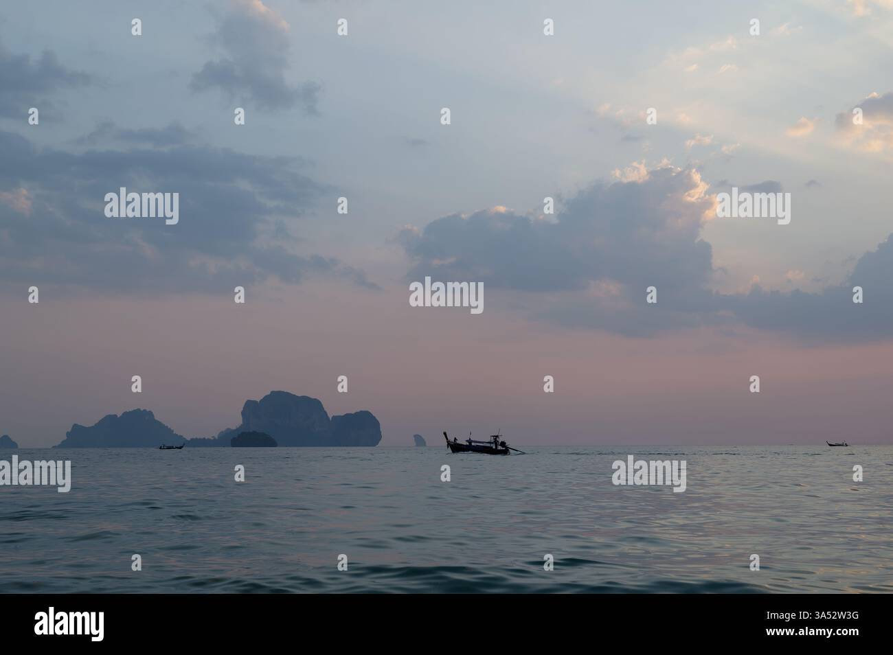 Una barca a coda lunga si allontana sulle acque calme al crepuscolo, con lontane isole calcaree che si stagliano contro un cielo pastello Foto Stock