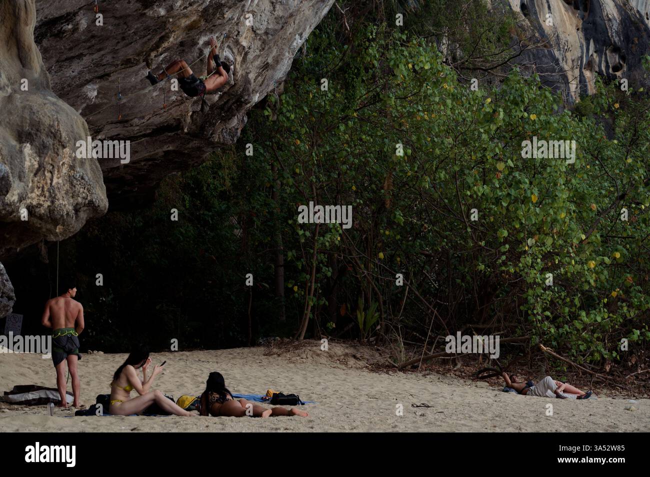 L'arrampicatore sale su una ripida sporgenza, mentre gli altri si rilassano sulla spiaggia sabbiosa sotto la scogliera coperta dalla giungla Foto Stock