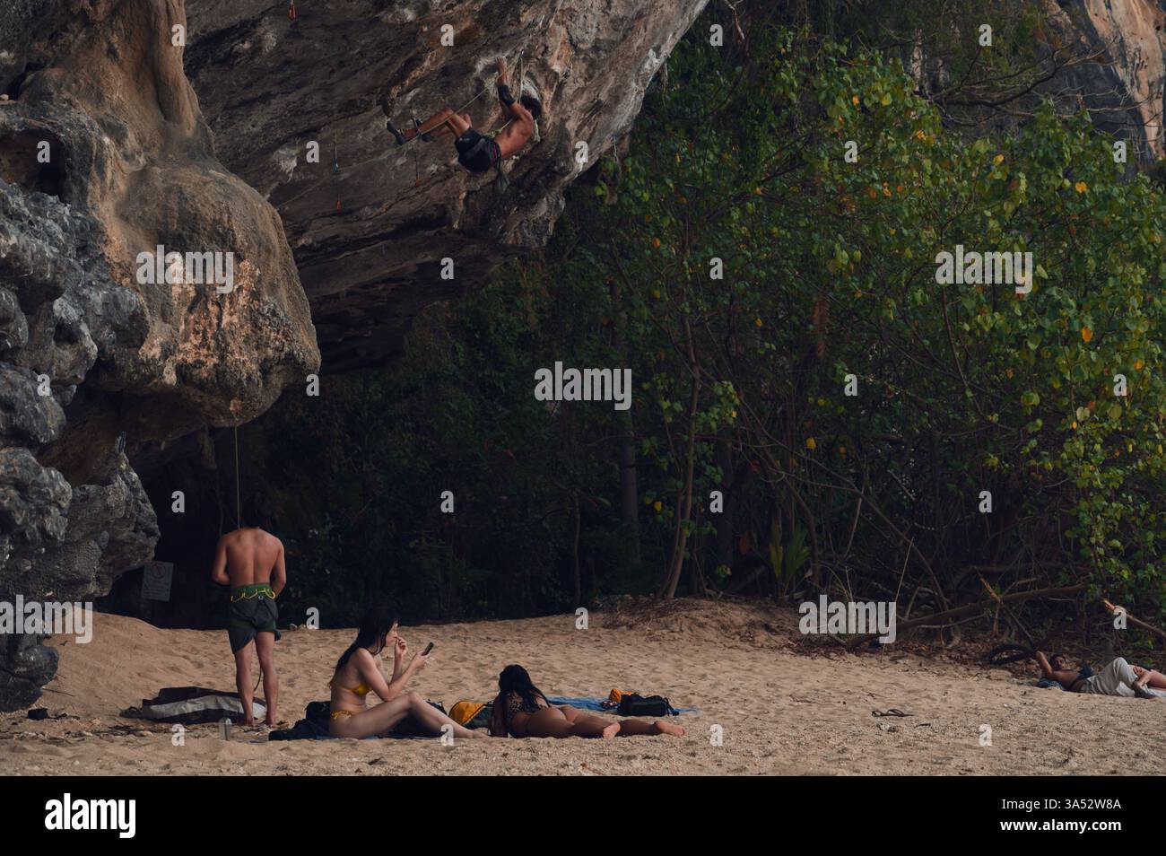 L'arrampicatore sale su una parete calcarea mentre un addetto alla cintura gestisce la corda. Nelle vicinanze, due amanti della spiaggia si rilassano sulla spiaggia Foto Stock
