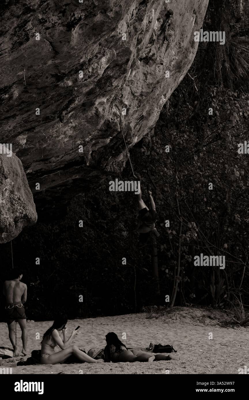 L'arrampicatore di roccia è sospeso a testa in giù da una corda su una sporgenza di pietra calcarea mentre gli amanti della spiaggia si rilassano sulla sabbia sottostante Foto Stock