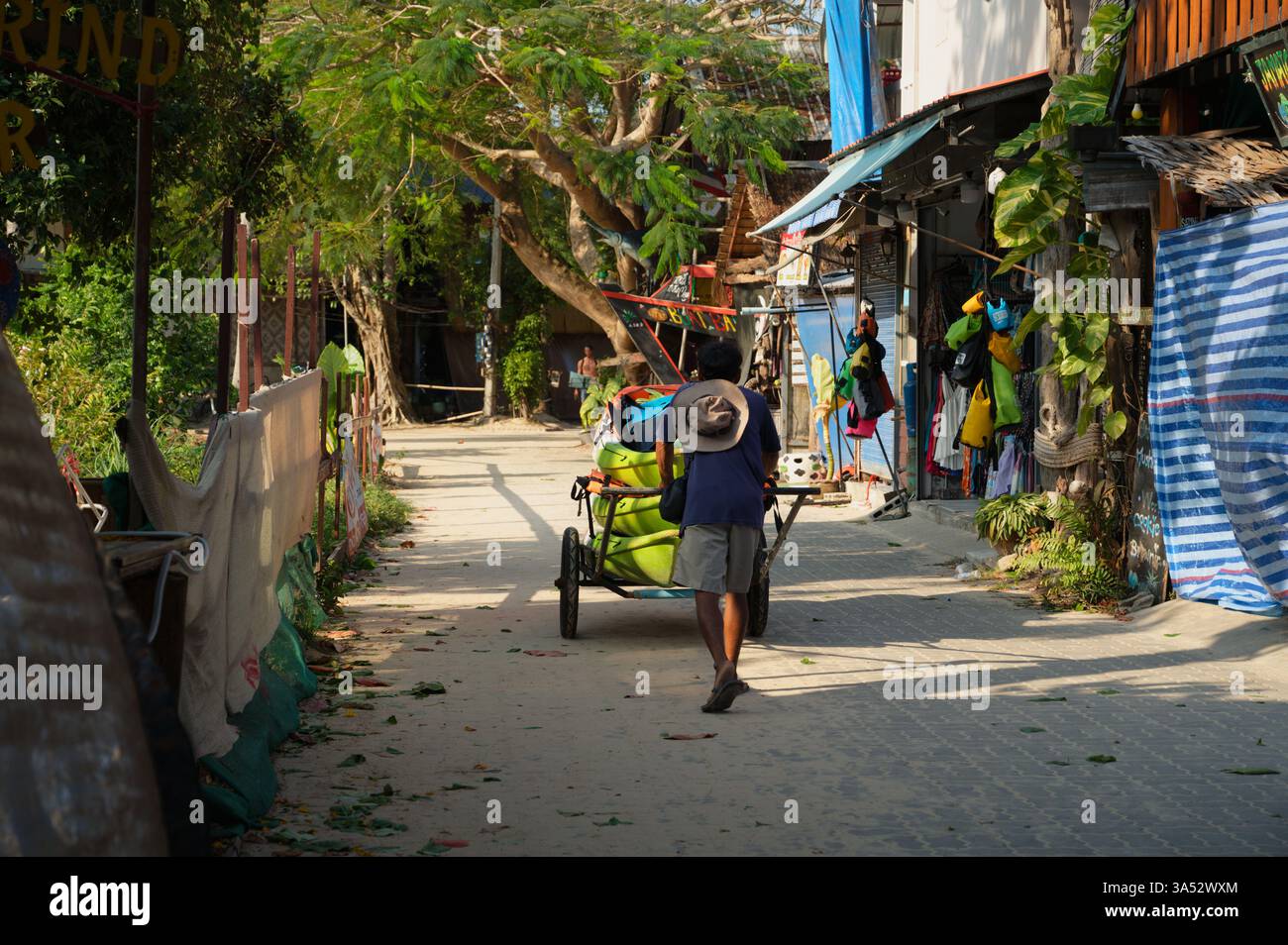 L'uomo spinge un carro carico di kayak attraverso un percorso illuminato dal sole a Railay, durante la mattina, passando per negozi chiusi Foto Stock