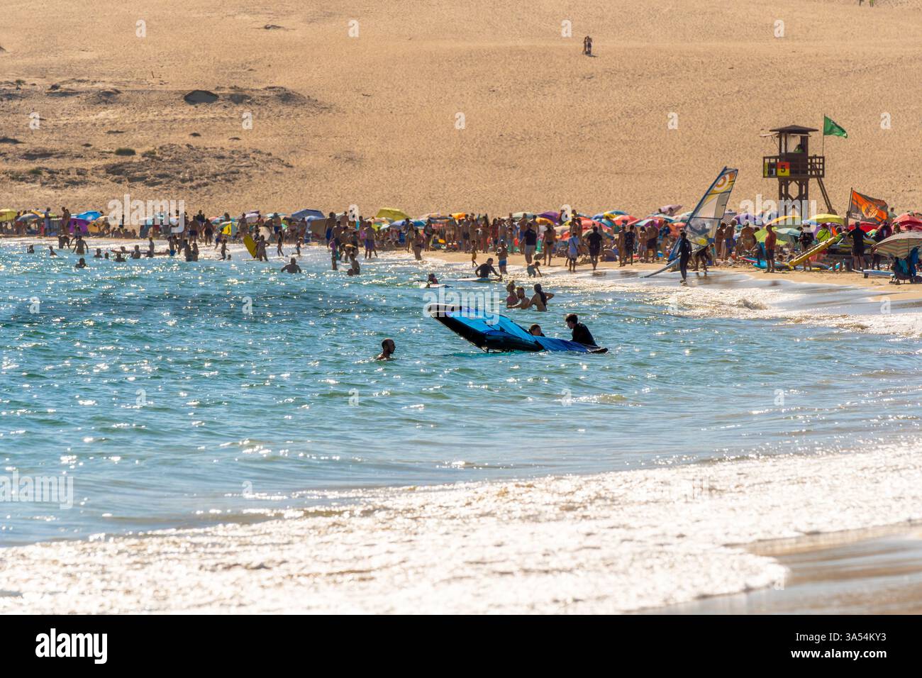 Spiaggia piena di persone e studenti della scuola di kitesurf che entrano in riva al mare con il sole del tramonto riflesso nelle sue acque e le dune nel b Foto Stock
