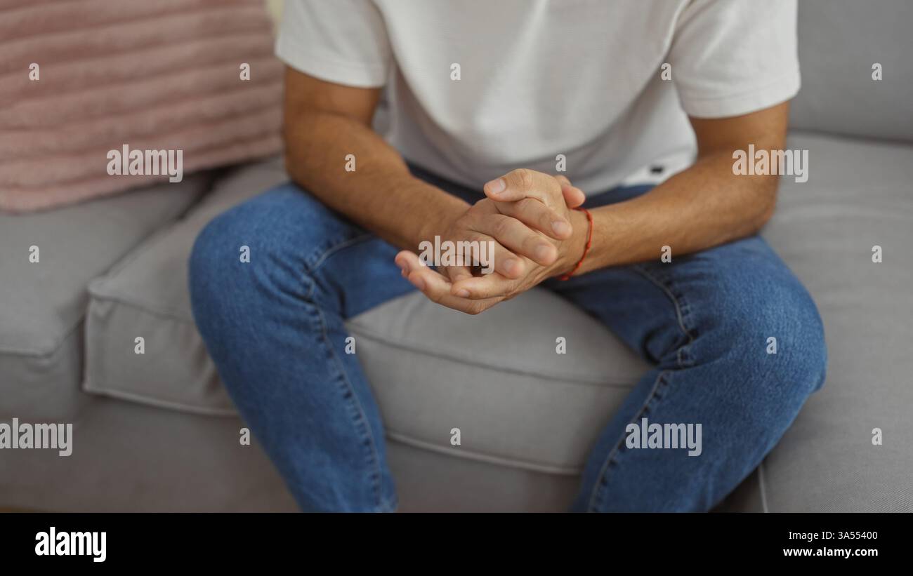 Giovane ispanico seduto in casa con le mani strette, vestito in modo informale con jeans e camicia bianca, creando un'atmosfera rilassata e premurosa a casa Foto Stock