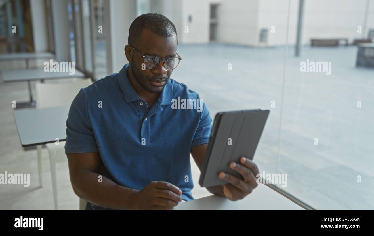 Giovane uomo dall'espressione seria che guarda un tablet in un ambiente di ufficio moderno, circondato da luce naturale e arredamento minimalista, si impegna in un ambiente di lavoro mirato Foto Stock