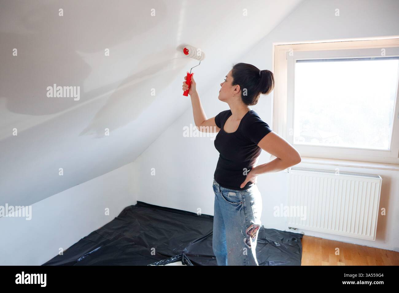 Donna con una t-shirt nera che dipinge un muro bianco con un rullo Foto Stock