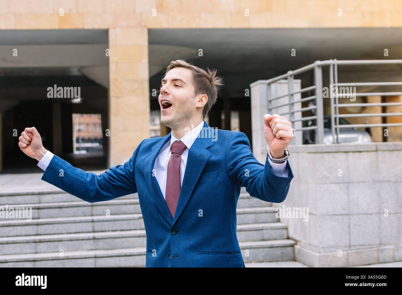 Allegro uomo d'affari con abito blu e cravatta rossa all'esterno con le braccia alzate, che esprime eccitazione e celebrazione, simboleggiando l'acconciatura professionale Foto Stock